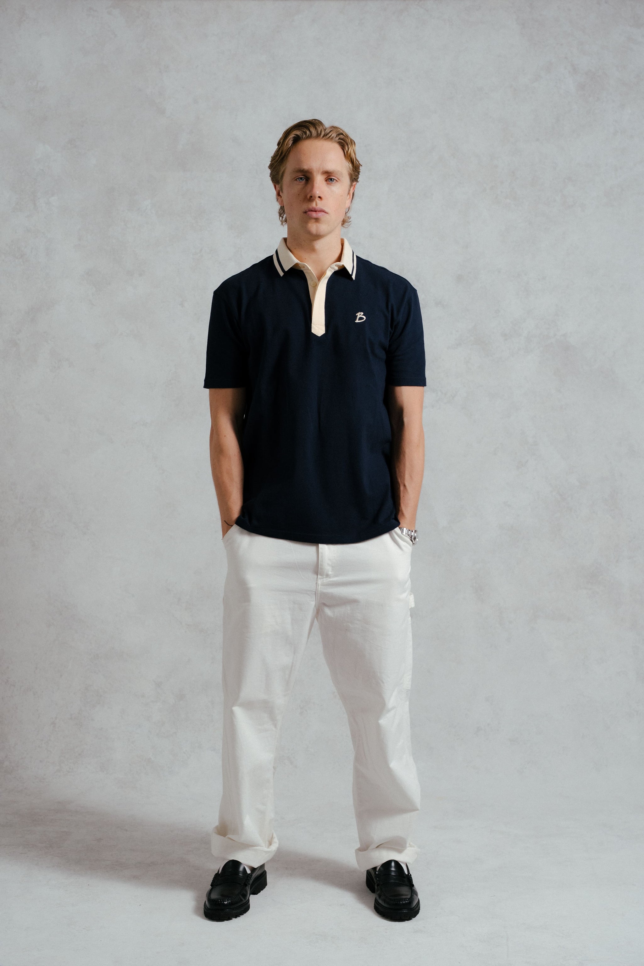 Man wearing a navy polo shirt with a white collar and white pants against a plain background