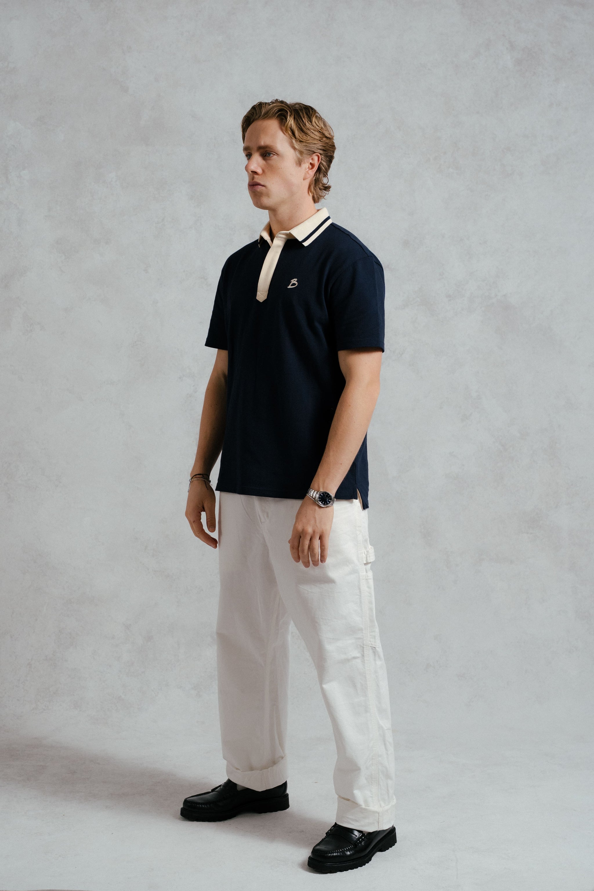 Man wearing a navy polo shirt with a white collar, white pants, and black shoes on a plain background
