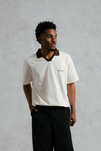 Man wearing a white polo shirt with a collar and black pants against a gray background